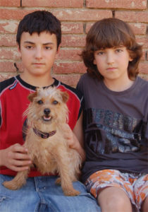 Dos nois i un gos davant d'un mur de maons.